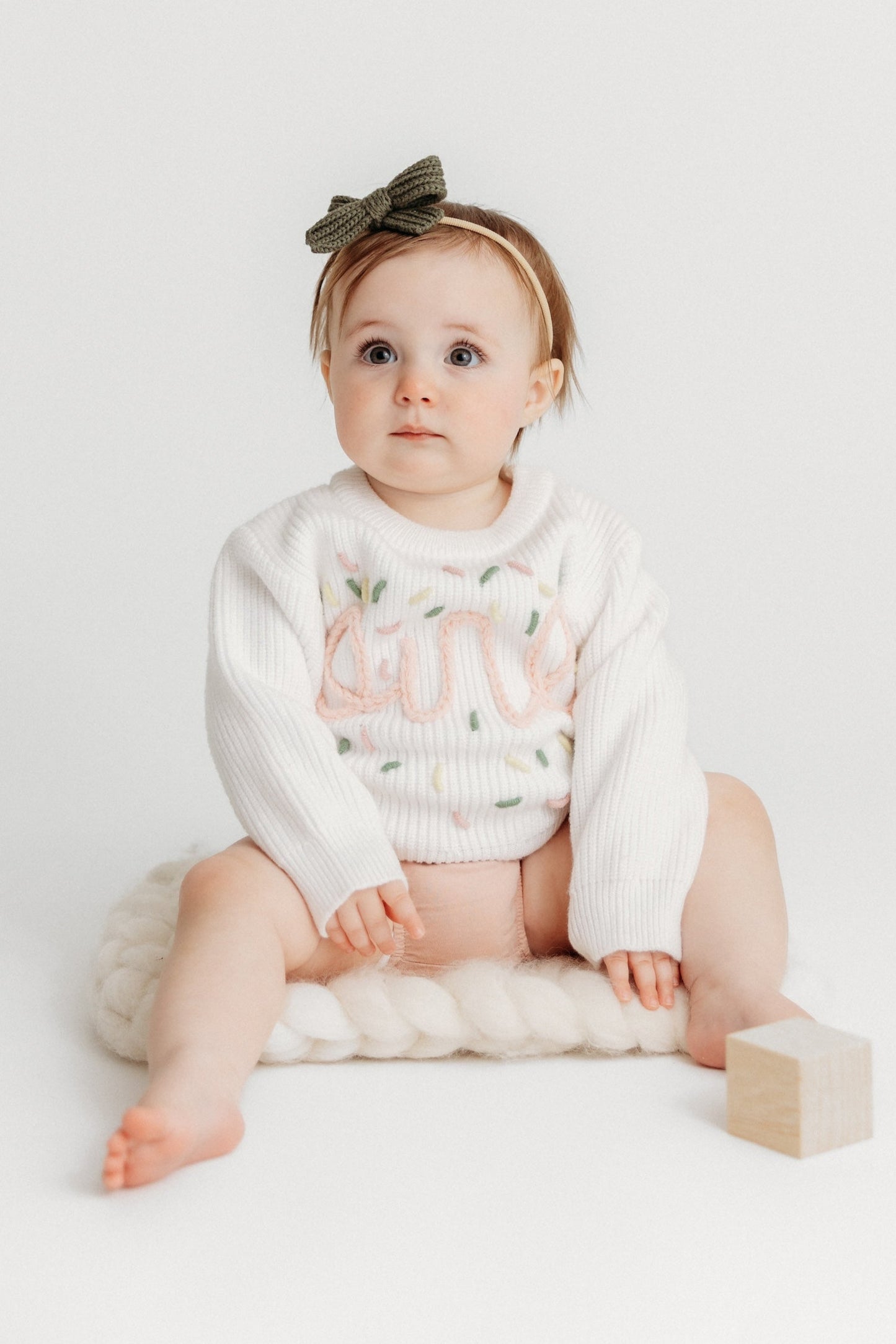 Baby sitting on a white surface with a white outfit and bow headband.