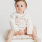 Baby sitting on a white surface with a white outfit and bow headband.