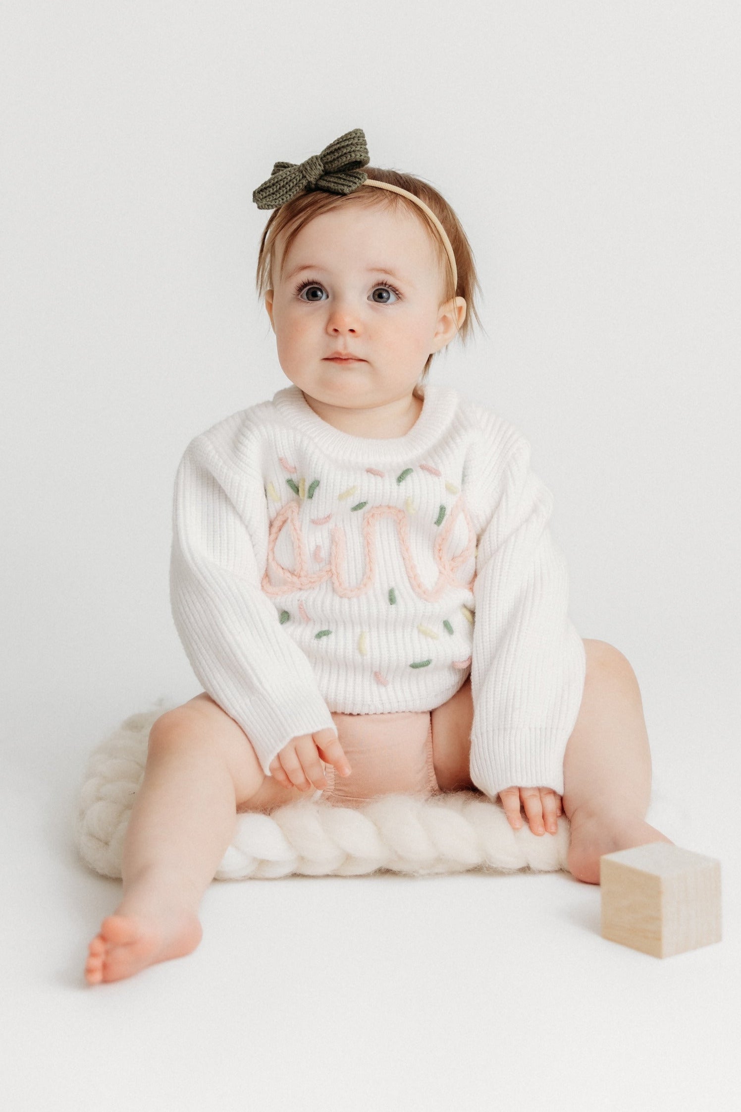 Baby sitting on a white surface with a white outfit and bow headband.