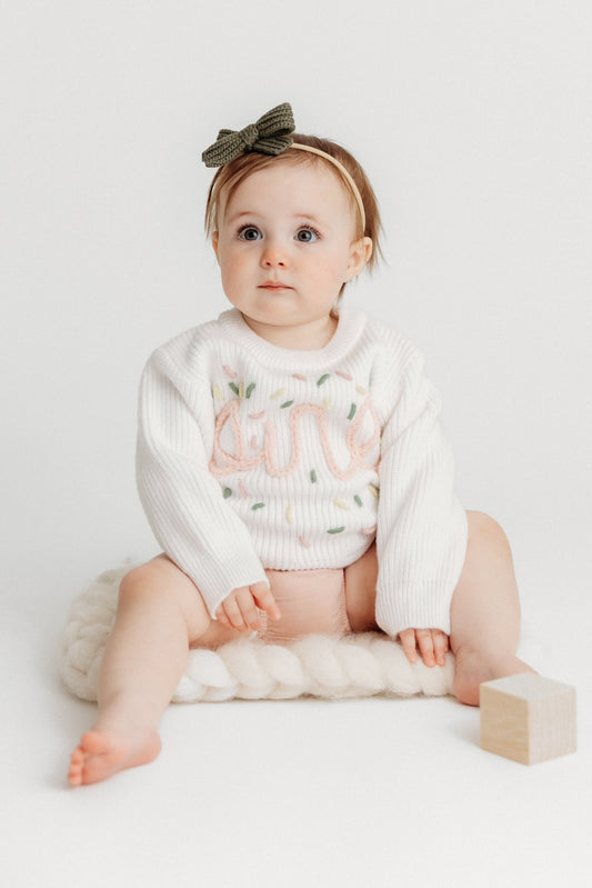 Baby sitting on a white surface with a white outfit and bow headband.